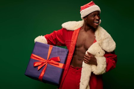 hombre americano africano feliz en traje de Navidad en cuerpo sin camisa sosteniendo regalo enorme en verde