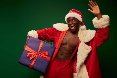 african american guy in christmas costume on shirtless body holding present and waving hand on green