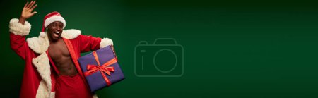 Photo for Hot african american guy in santa claus costume holding present and waving hand on green, banner - Royalty Free Image