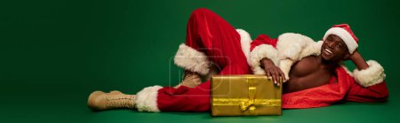 souriant sexy afro-américain en costume de Père Noël couché près de boîte cadeau brillant sur vert, bannière