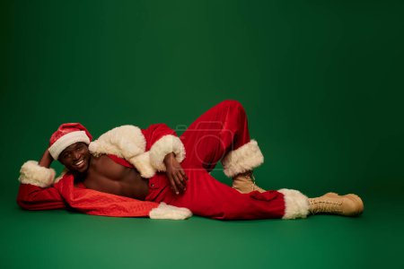 heißer afrikanisch-amerikanischer Kerl im Weihnachtsmann-Kostüm auf hemdslosem Körper liegend und lächelnd auf grünem Grund