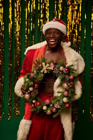 hombre afroamericano complacido en traje de santa con corona de Navidad cerca de oropel de oro en verde