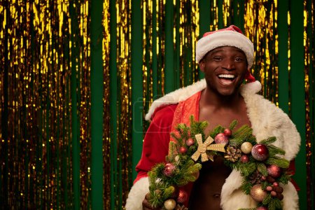 laughing african american guy in santa costume with christmas wreath near shiny decor on green