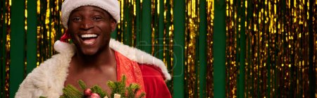 Foto de Despreocupado hombre afroamericano en traje de Navidad riendo cerca de oropel de oro en verde, bandera - Imagen libre de derechos