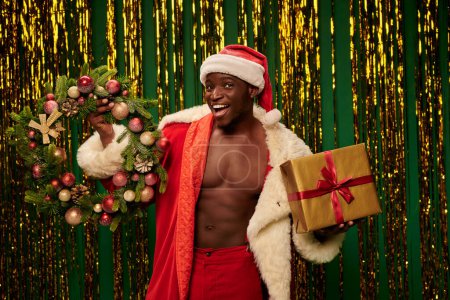 hombre afroamericano disfrazado de santa con corona de Navidad y caja de regalo con fondo de oropel dorado