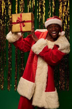 aufgeregter afrikanisch-amerikanischer Mann im Weihnachtskostüm zeigt Geschenk auf grünem und glänzendem Hintergrund