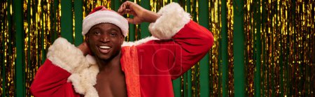 Photo for Jolly african american man in santa costume smiling at camera near golden tinsel on green, banner - Royalty Free Image