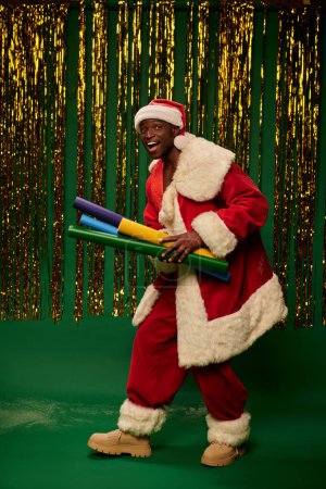 joyeux homme afro-américain en costume de Père Noël avec des rouleaux de papier coloré sur fond vert avec tinsel