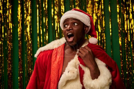erstaunt afrikanisch-amerikanischer Mann im Weihnachtsmannkostüm mit offenem Mund und Weihnachtstasche neben goldenem Lametta
