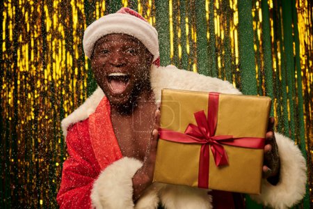 homme afro-américain énergisé en costume de Père Noël avec sac de Noël et bouche ouverte sous la neige