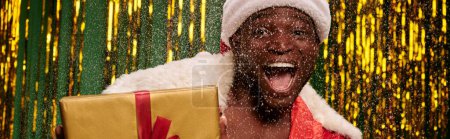 erstaunt afrikanisch-amerikanischer Mann im Weihnachtsmannkostüm mit Geschenkbox im schneebedeckten und glänzenden Atelier, Banner