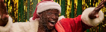 cheerful african american man in santa costume gesturing under snowfall on golden backdrop, banner