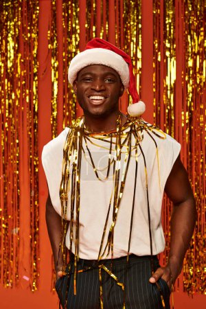 fröhlicher afrikanisch-amerikanischer Mann mit Weihnachtsmütze und weißem Tanktop in der Nähe von goldenem Lametta auf rotem Hintergrund