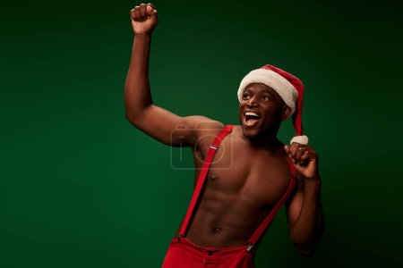 emocionado hombre afroamericano sin camisa en sombrero de Navidad y pantalones regocijándose y gritando en verde