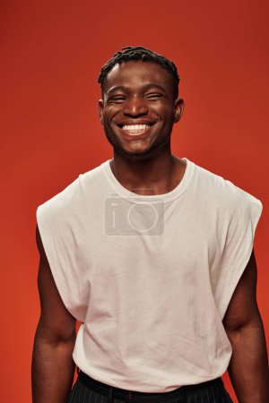 happy and sporty african american man in white tank top smiling at camera on red, modern male model