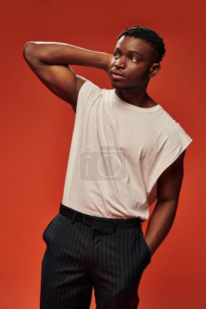 Téléchargez les photos : Jeune homme afro-américain en tenue décontractée tendance tenant la main dans la poche et regardant loin sur le rouge - en image libre de droit