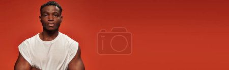 confident dark skinned man in white tank top looking at camera on red studio backdrop, banner