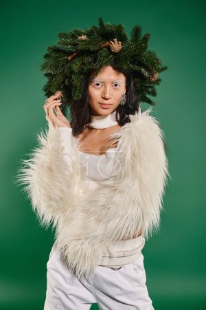 Winterkonzept, asiatische Frau mit weißem Make-up und trendigem Outfit posiert im Kranz vor grünem Hintergrund