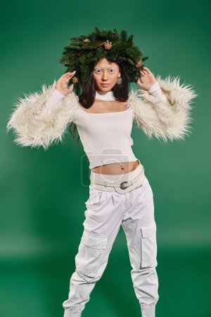 Winterkonzept, hübsche Frau mit weißem Make-up und trendigem Outfit posiert im Kranz vor grünem Hintergrund