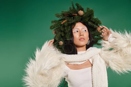 holiday spirit, brunette asian woman with white makeup and trendy outfit posing in wreath on green
