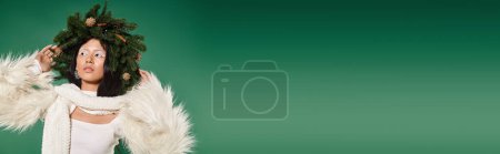 holiday banner, brunette asian woman with white makeup and trendy outfit posing in wreath on green