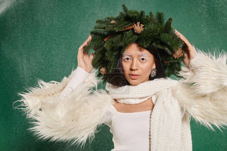 winter time, beautiful woman with white makeup and trendy outfit posing in wreath under falling snow