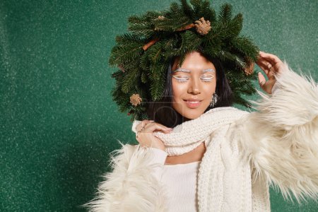 winter time, happy woman with white makeup and trendy outfit adjusting wreath under falling snow