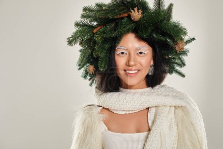 winter concept, happy asian woman with natural pine wreath posing in white clothes on grey backdrop
