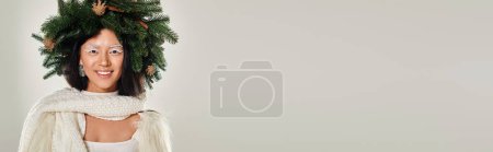 winter banner, happy asian woman with natural pine wreath posing in white clothes on grey backdrop