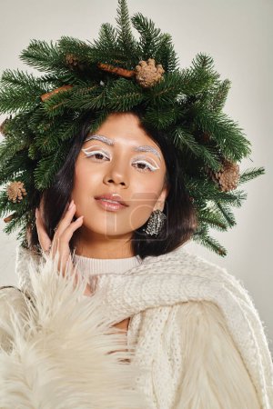 belleza de invierno, mujer atractiva con corona de pino verde posando en ropa blanca sobre fondo gris