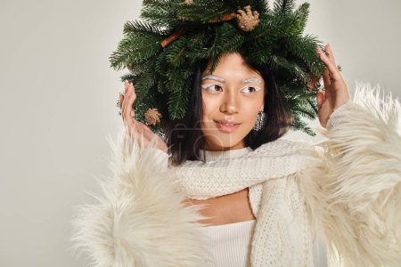belleza de invierno, mujer positiva con corona de pino natural posando en ropa blanca sobre fondo gris