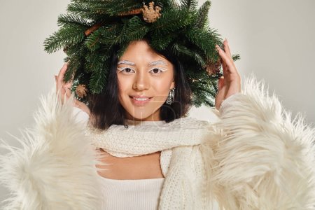 winter beauty, joyful asian woman with natural pine wreath posing in white clothes on grey backdrop