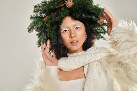 winter beauty, enchanted woman with natural pine wreath posing in white clothes on grey backdrop