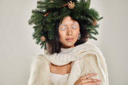 winter beauty, charming woman with natural pine wreath posing in white clothes on grey backdrop
