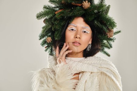 asian beauty, alluring woman with natural pine wreath posing in white clothes on grey backdrop