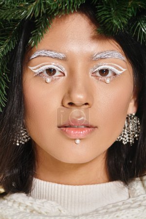 winter beauty, close up of attractive woman with white eye makeup and beads on face posing in wreath