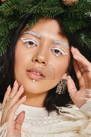winter trends, asian woman with white eye makeup and beads on face posing in wreath, look at camera