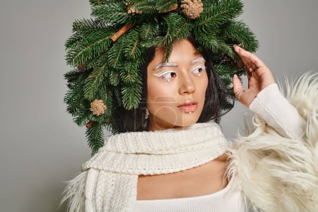 winter trends, beautiful woman with white eye makeup and beads on face posing in wreath on grey