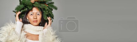 winter banner, attractive woman with white eye makeup and beads on face posing in wreath on grey