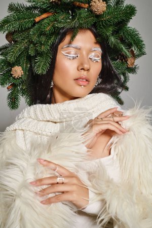 holiday style, attractive woman with white eye makeup and beads on face posing in wreath on grey