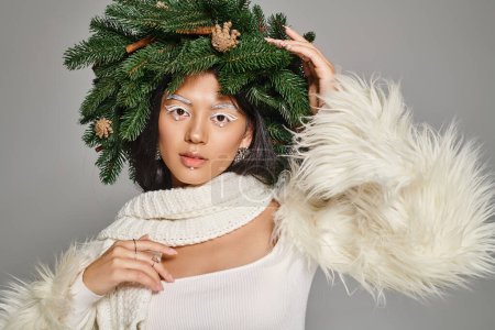 holiday style, charming woman with white eye makeup and beads on face posing in wreath on grey