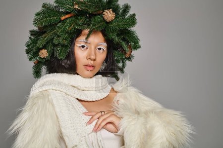 Urlaub Stil, asiatische Winterkönigin mit weißen Augen Make-up und Perlen im Gesicht posiert in Kranz auf grau