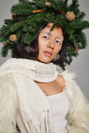 beautiful winter queen with white eye makeup and beads on face posing in wreath on grey background