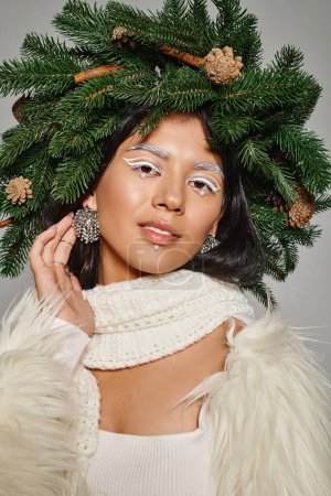 attractive woman with white eye makeup and beads on face posing in wreath and earrings on grey