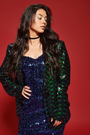 brunette asian woman in green dress and jacket with sequins posing with hand on hip on red backdrop