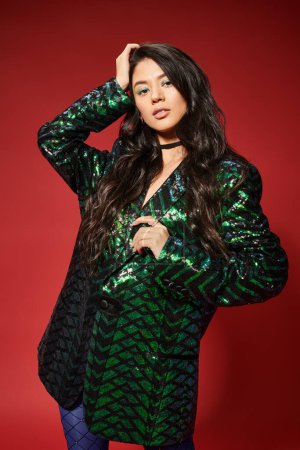 asian brunette young woman in trendy green jacket with sequins looking at camera on red backdrop