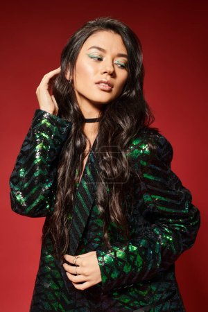 asian brunette woman in trendy green jacket with sequins adjusting wavy hair on red background