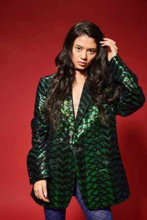 beautiful asian woman in trendy green jacket with sequins adjusting wavy hair on red background