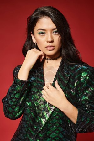 schöne asiatische Frau in stilvoller grüner Jacke mit Pailletten blickt in die Kamera auf rotem Hintergrund