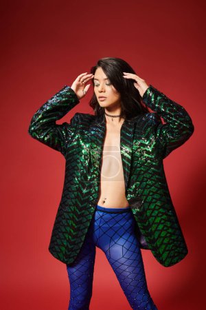 Téléchargez les photos : Sexy asiatique femme dans élégant vert veste avec sequins et bleu collants posant sur fond rouge - en image libre de droit